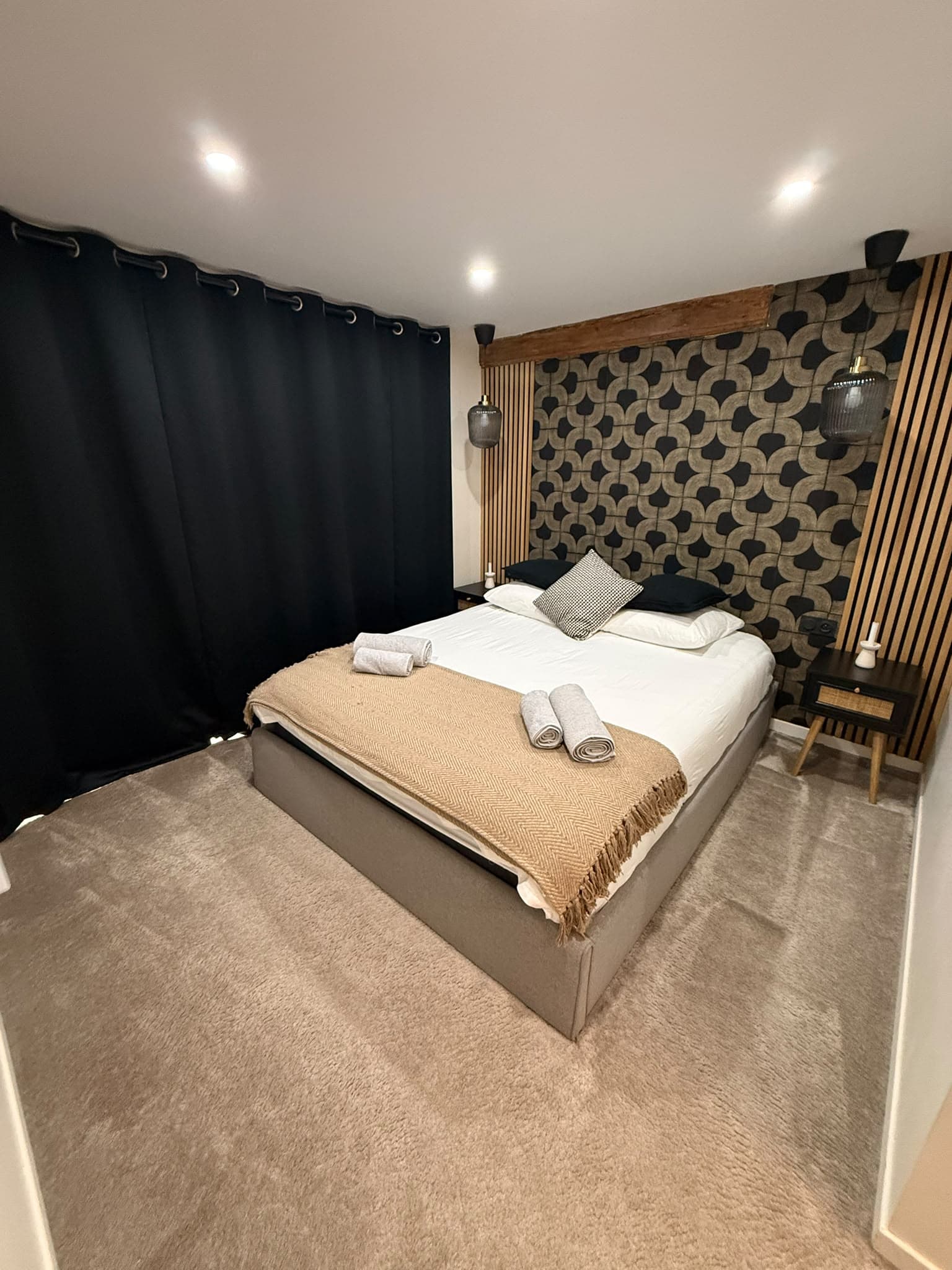 Chambre moderne avec lit double, jeté beige, rideaux noirs et mur à motifs géométriques.