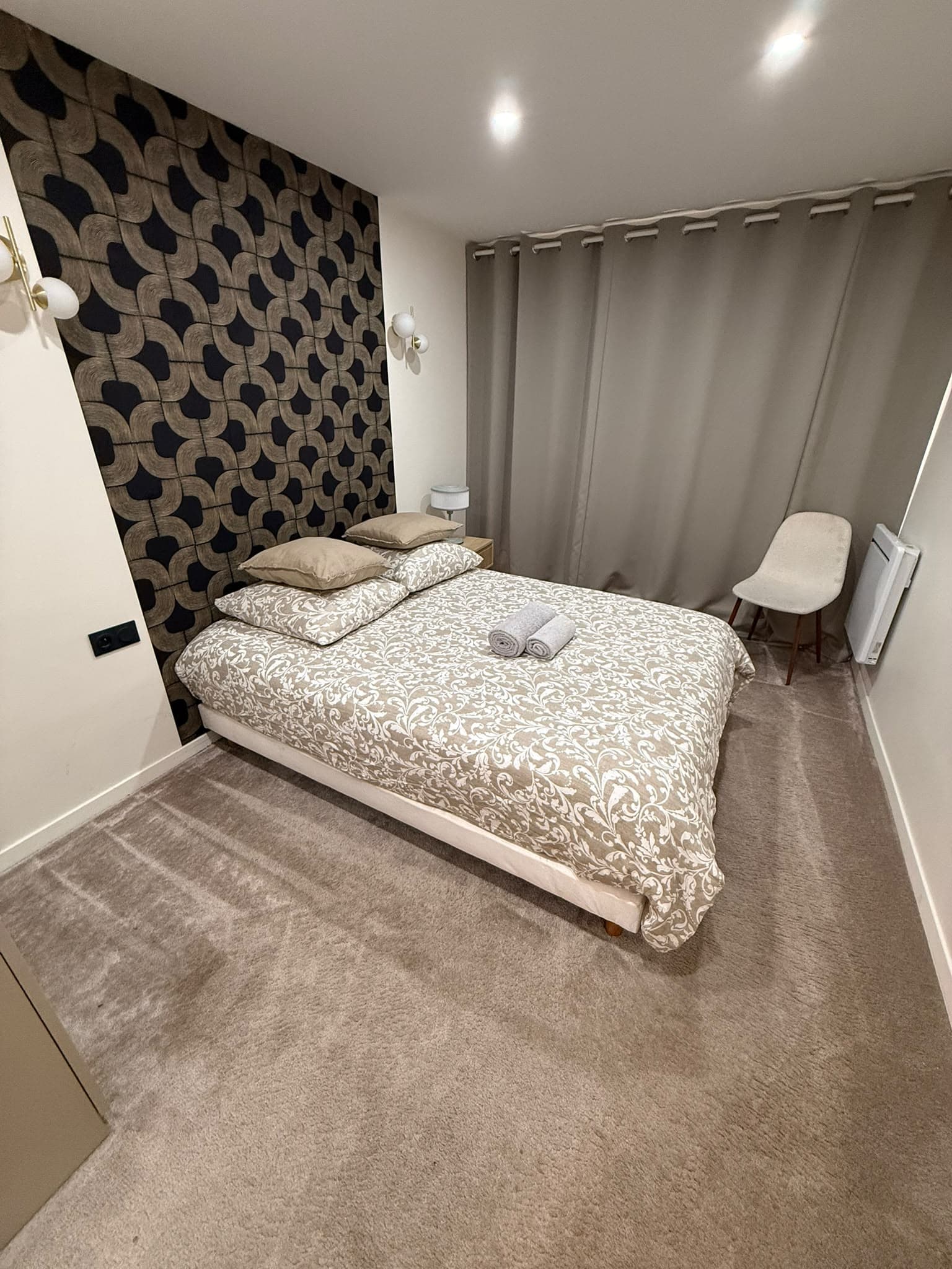 Chambre moderne avec lit double, couette fleurie, mur à motifs géométriques et rideaux beiges.