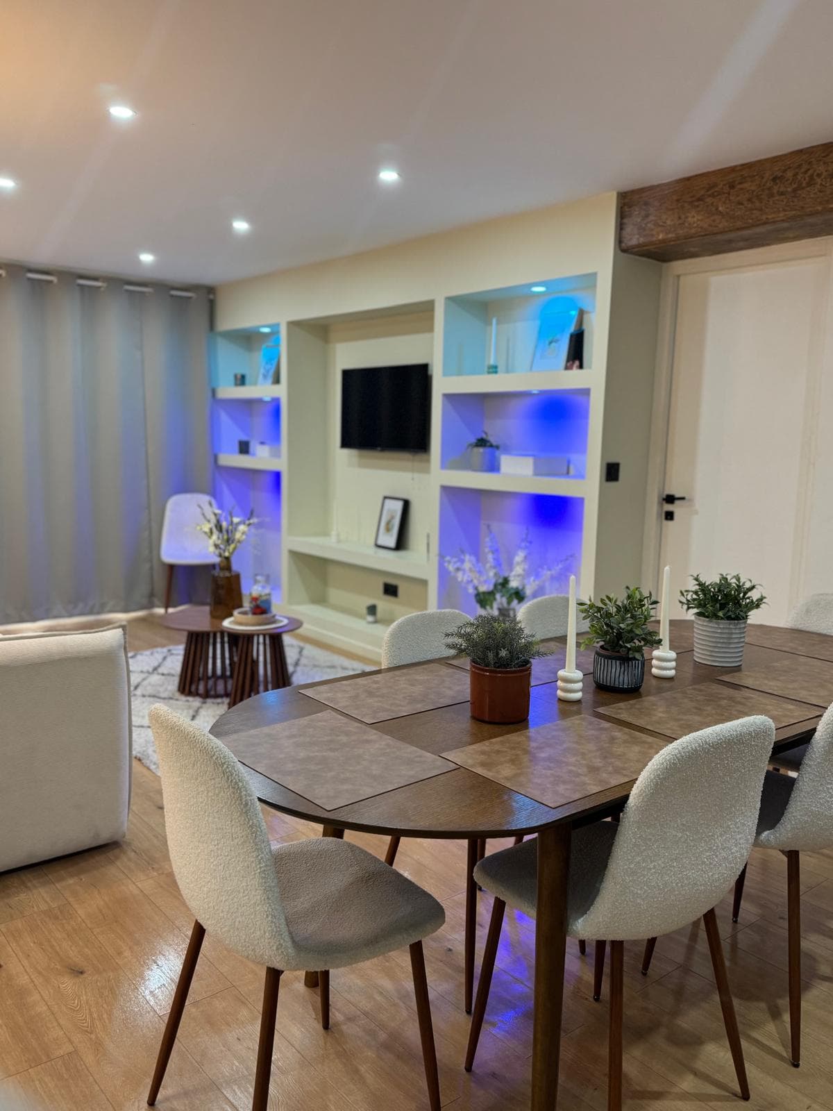 Table à manger ovale et chaises blanches devant un salon aux étagères éclairées en bleu.